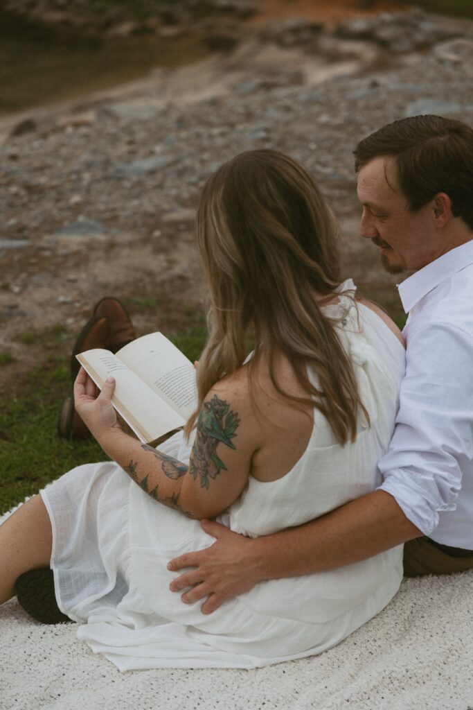 Man with his arm around a woman sitting on a blanket in the grass reading a book.