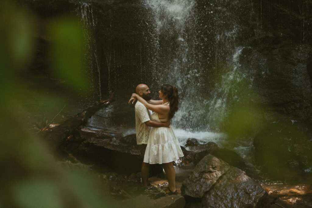 Man and woman hugging each other in front of waterfall with green leaves in foreground.