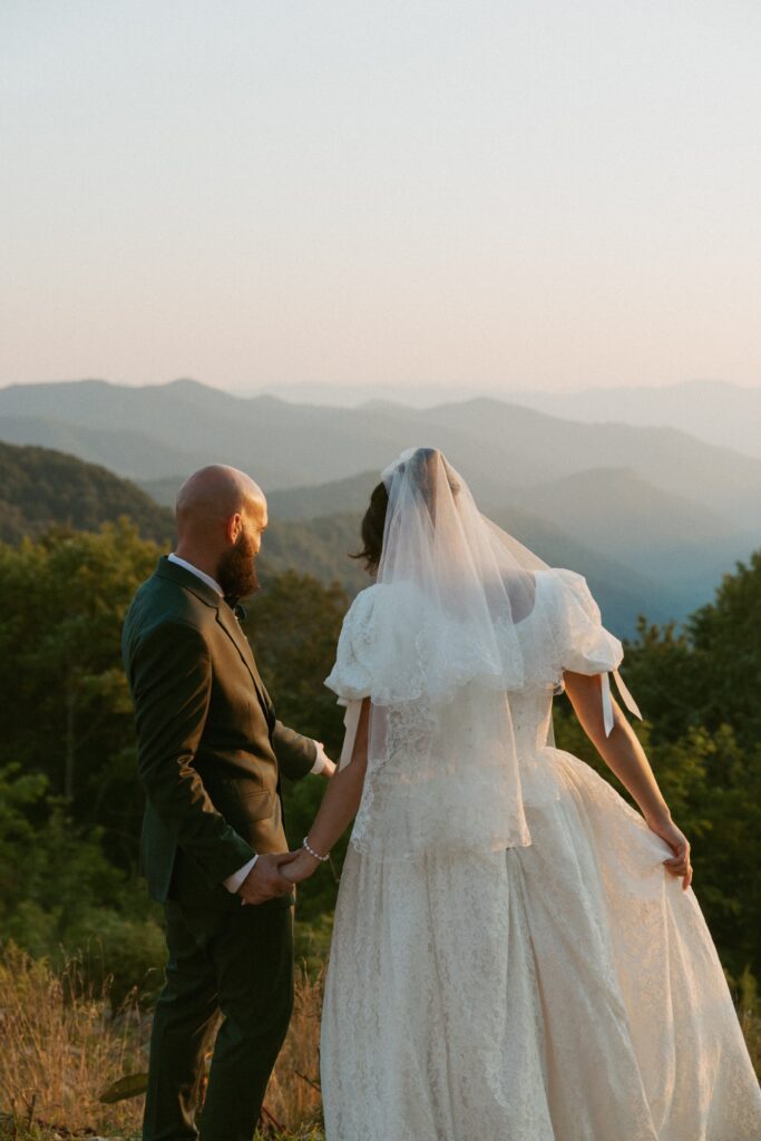 Man and woman in wedding attire holding hands and looking out at mountains during sunset.