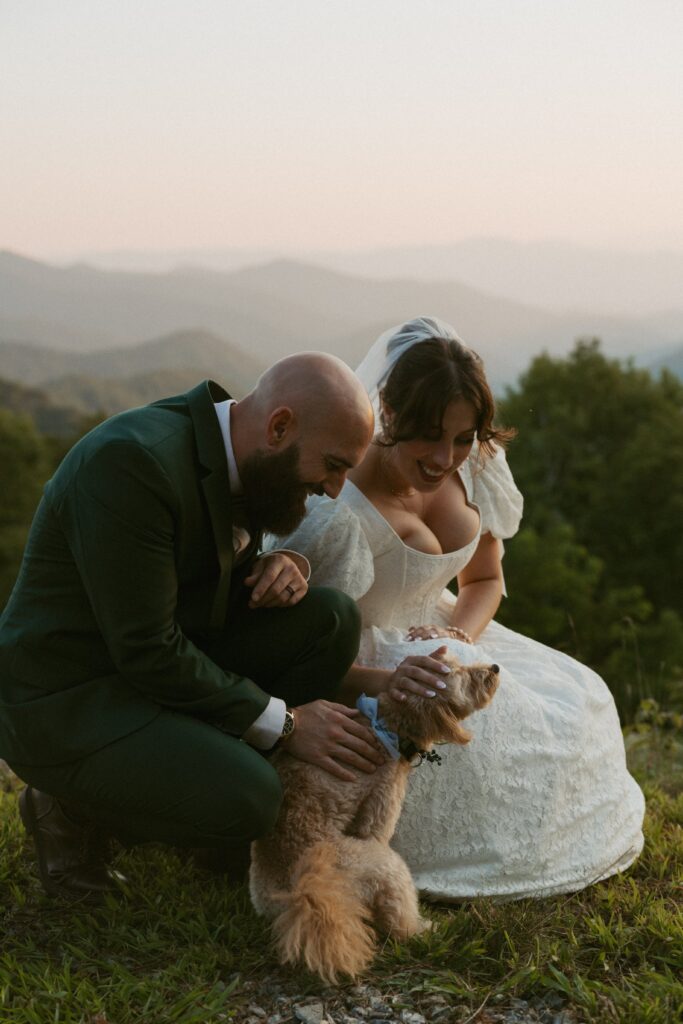 Man and woman in wedding attire playing with their dog in front of the mountains.