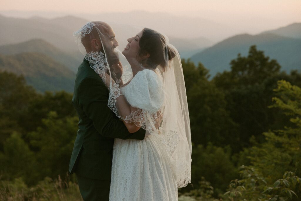 Man and woman laughing under brides veil on their wedding day with mountains behind them.