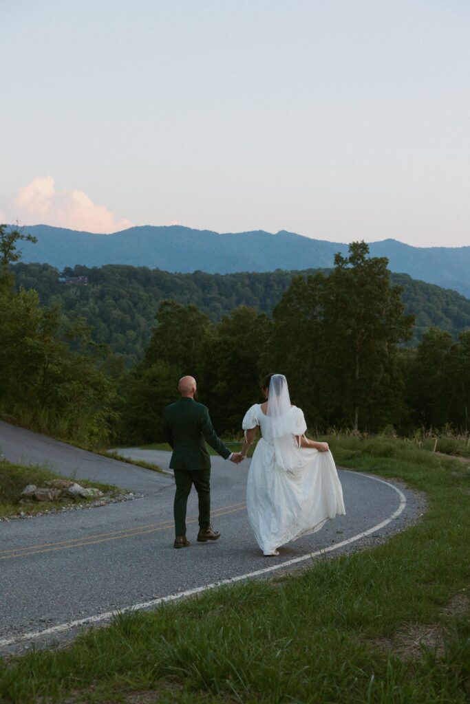 Man and woman in wedding attire holding hands and walking down road towards mountains.