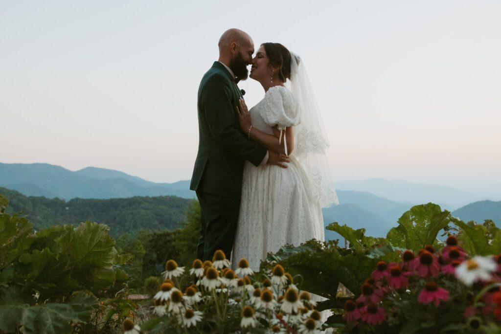 Man and woman in wedding attire smiling and going in for a kiss with mountains behind them and flowers in front of them.