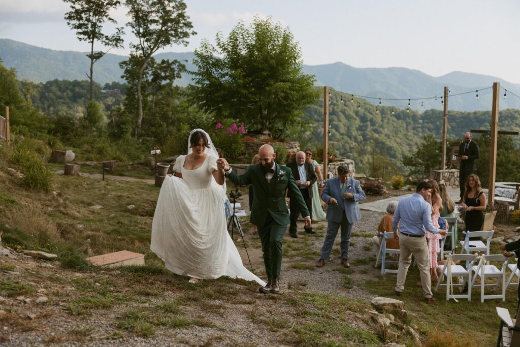 Man and woman walking away from their wedding ceremony with mountains and guests behind them.