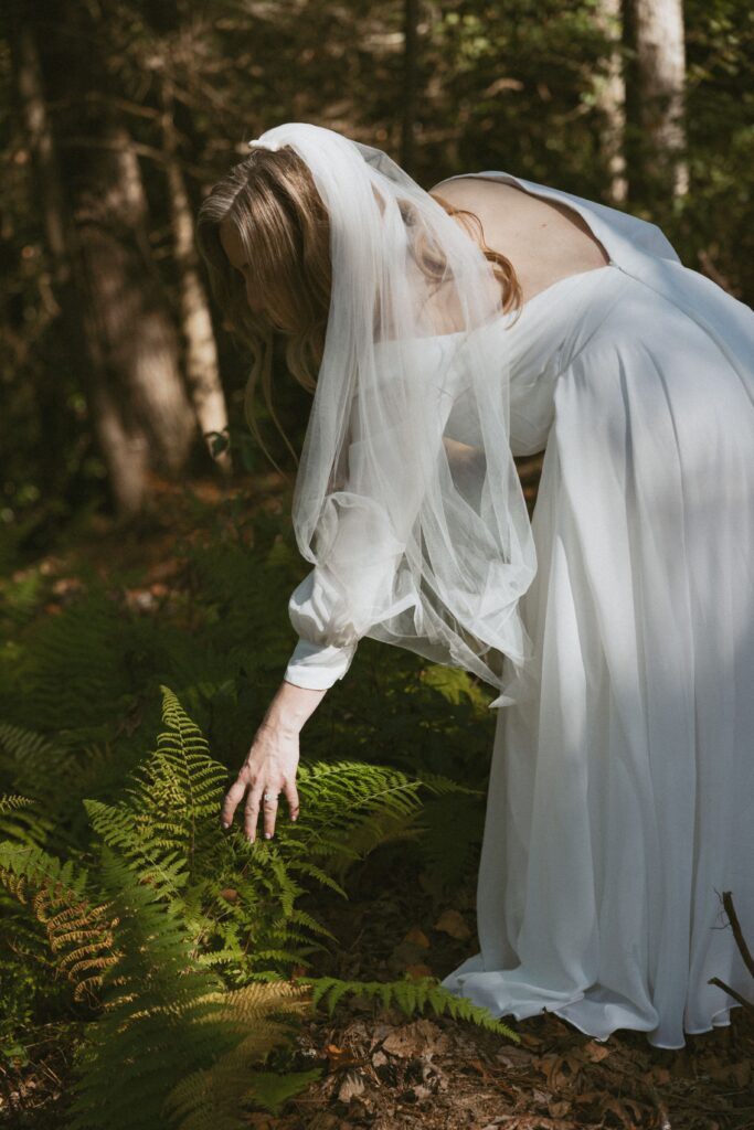 Woman in wedding dress bending down and touching ferns.