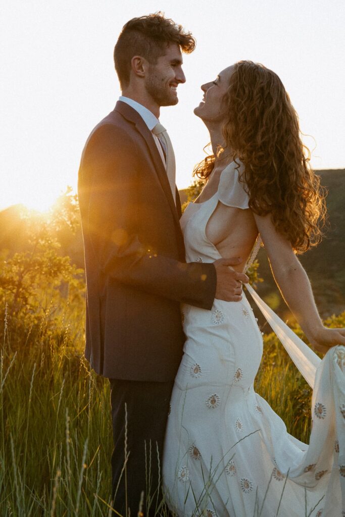 Man and woman laughing while in wedding attire with mountains and sunset behind them.