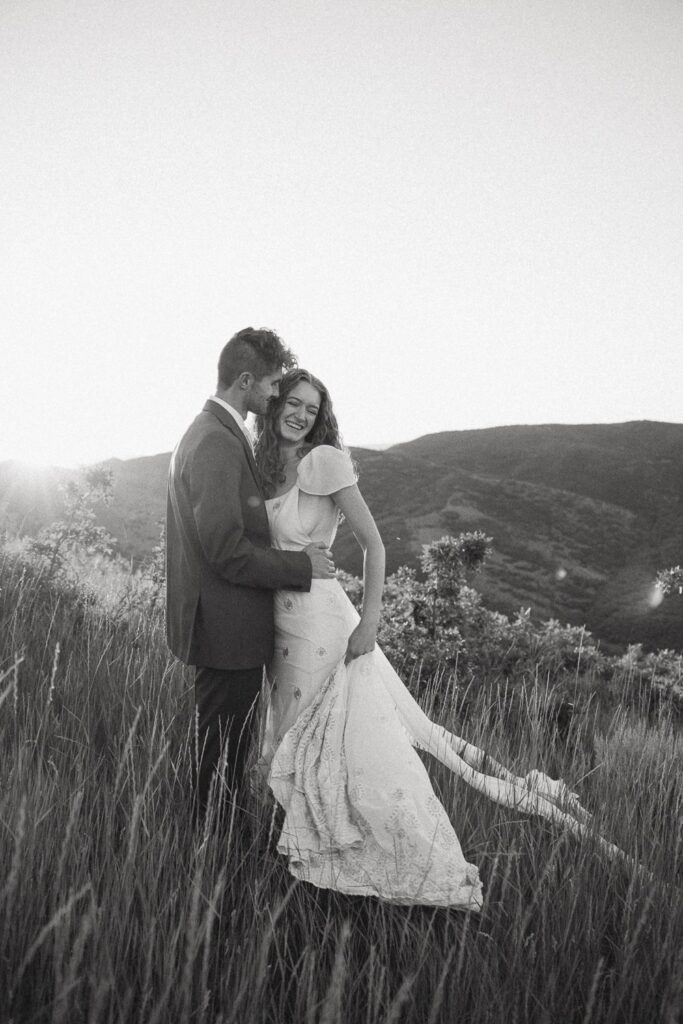 Black and white photo of man and woman laughing while in wedding attire with mountains and sunset behind them.