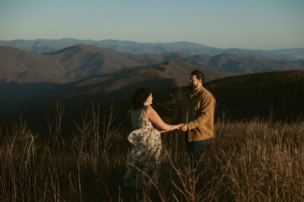 Man and woman holding hands and smiling at each other with mountains behind them.