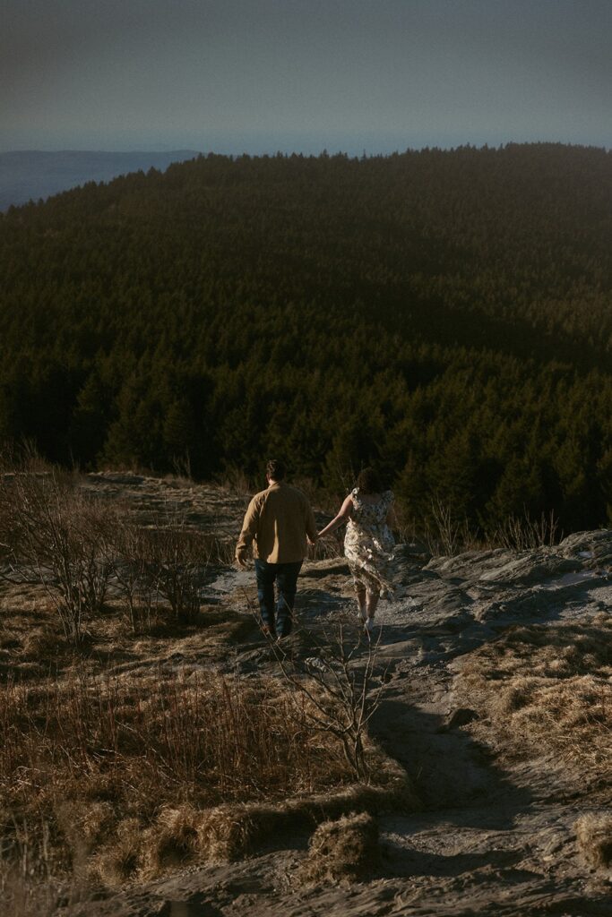 Man and woman walking down a trail while holding hands with trees and mountains in front of them.