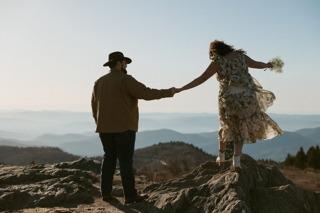 Man holding woman's hand while she tries to walk across a rock with the mountains in the background.