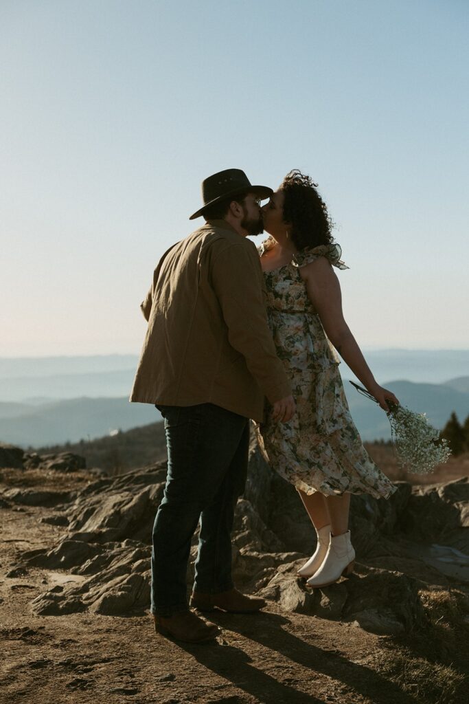 Woman standing on a rock and kissing a man with the mountains behind them.