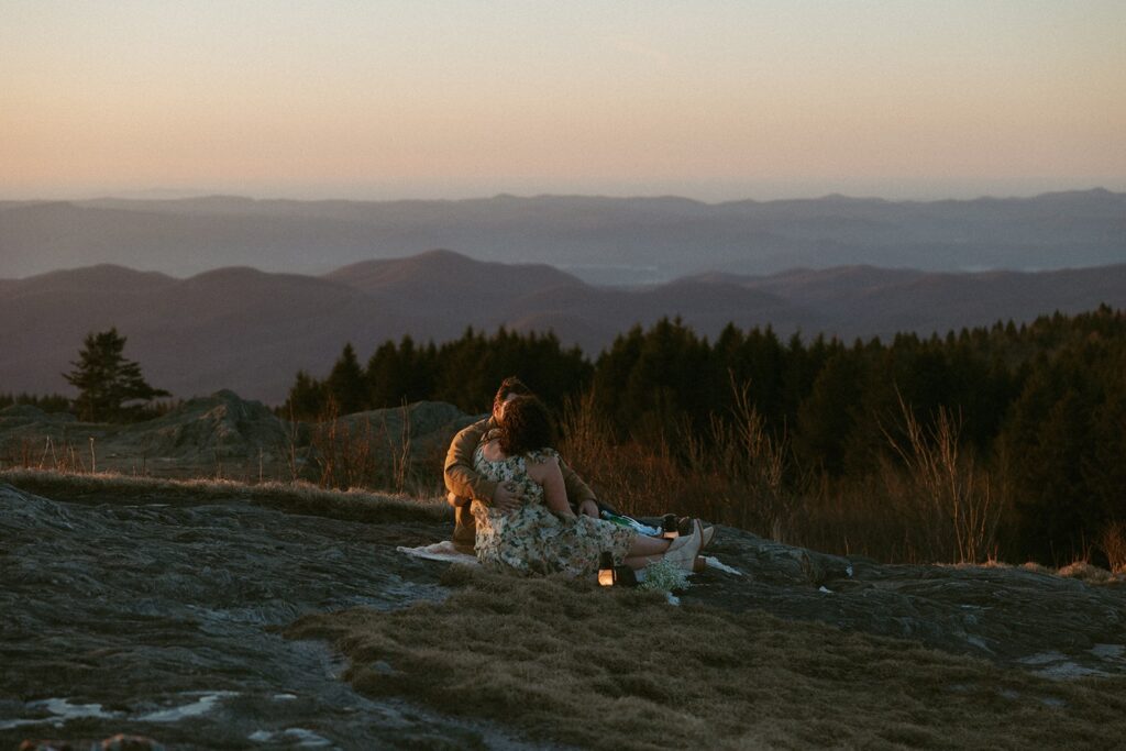 Man and woman sitting on a blanket and holding each other with mountains at sunrise behind them.