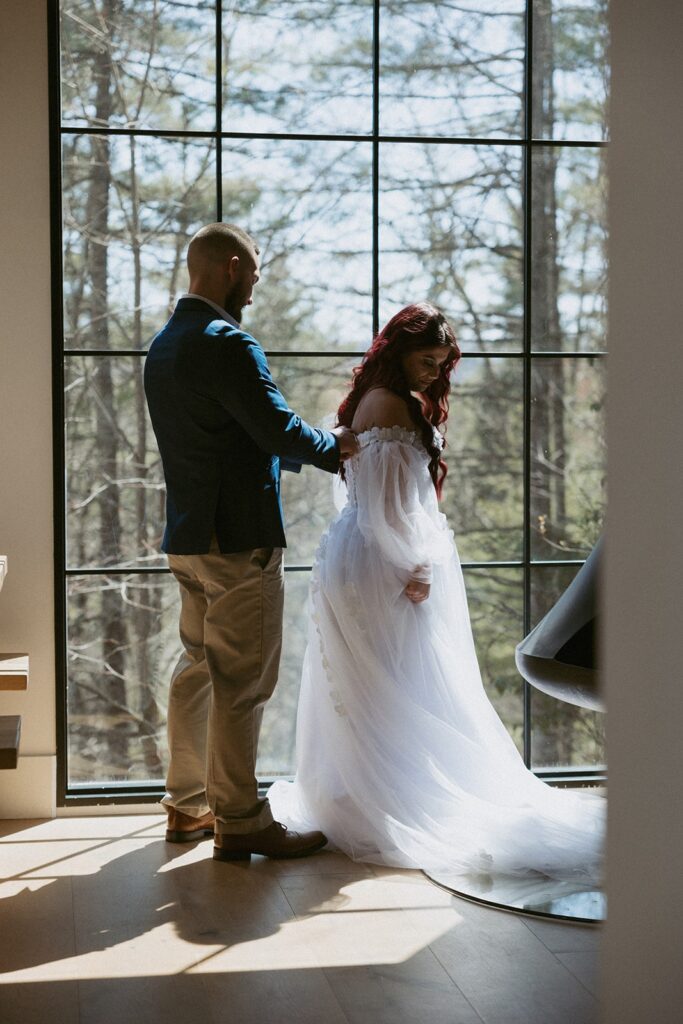 Man helping woman zip up wedding dress in front of window in airbnb.