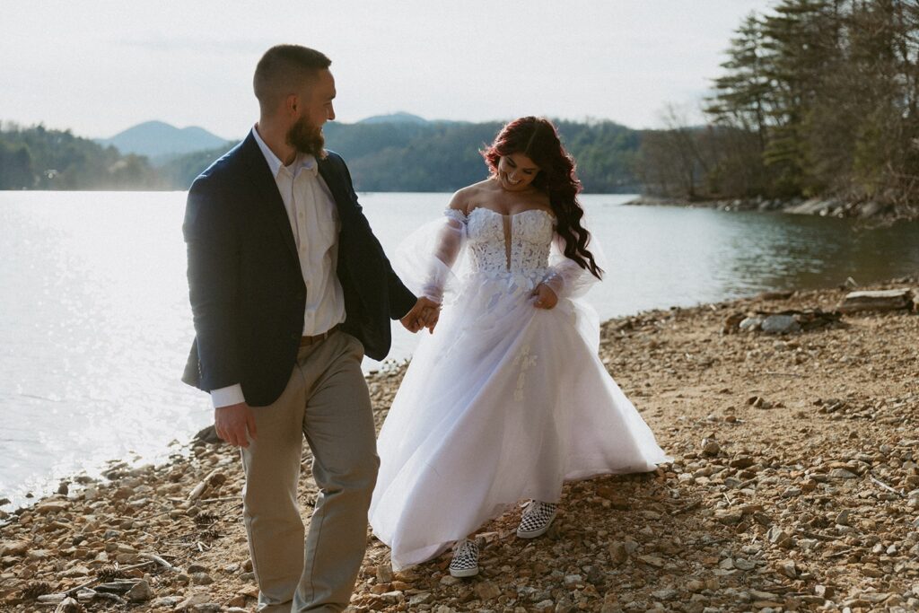 Man and woman in wedding attire walking across rocky lake shore with mountains behind them.