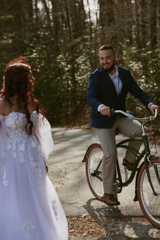 Man in suit riding a bike while woman in wedding dress watches him.