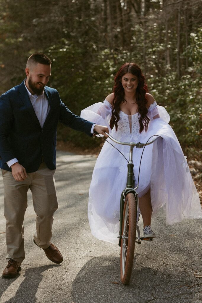 Woman in wedding dress riding a bike while man in suit helps her.