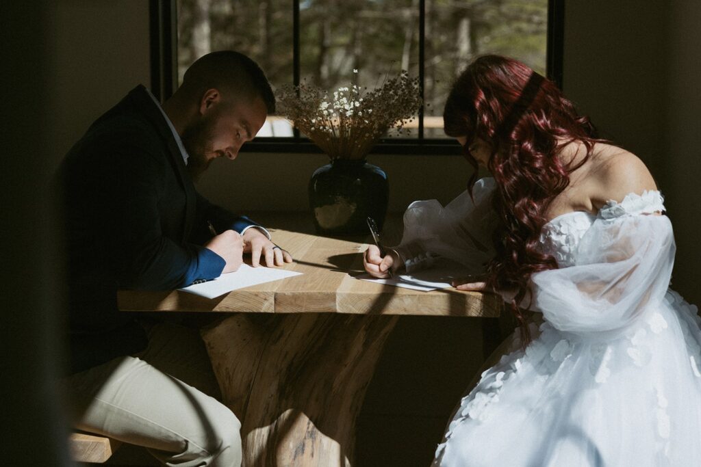 Man and woman in wedding attire sitting at table writing vows.