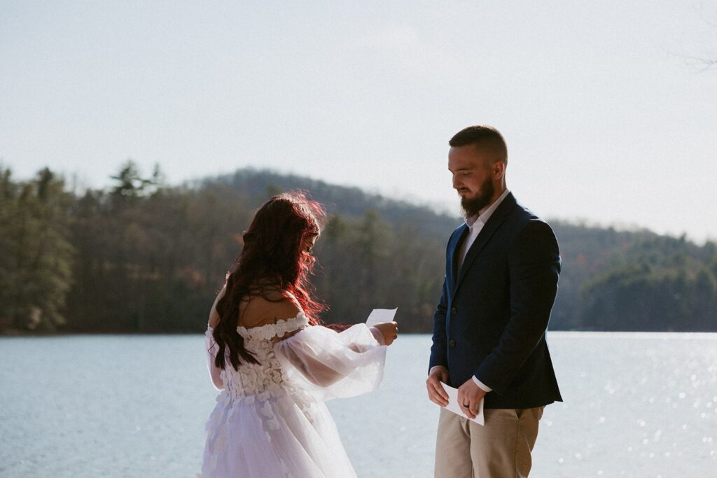 Man and woman in wedding attire reading notes to each other in front of lake and mountains.