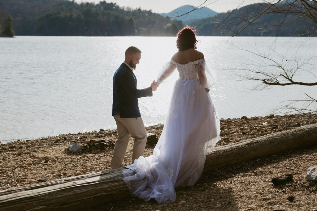 Man in suit helping woman in wedding dress walk across a log in front of lake and mountains.