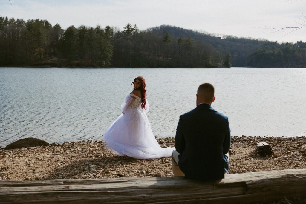 Man in suit sitting on log looking at woman in wedding dress with lake and mountains behind her.