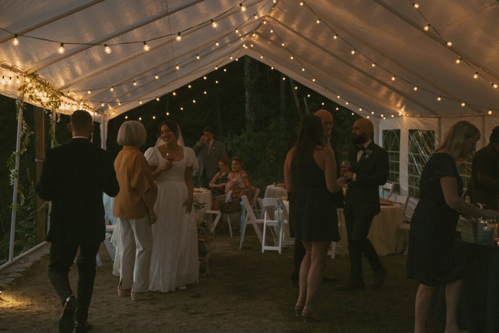 Bride and groom plus other guests sitting, drinking and talking under tent with twinkle lights at night.