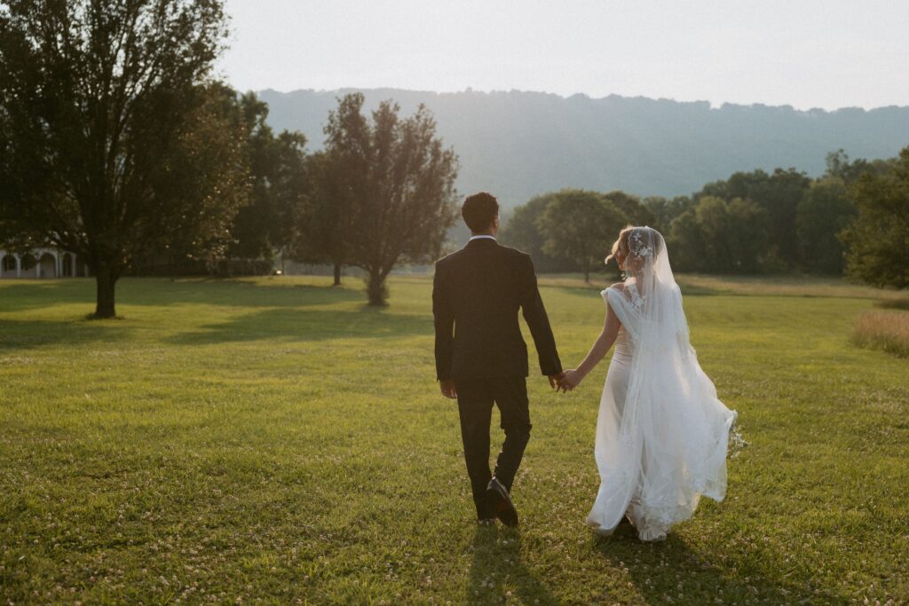 Bride and groom walking through field at sunset holding hands with mountains in front of them.