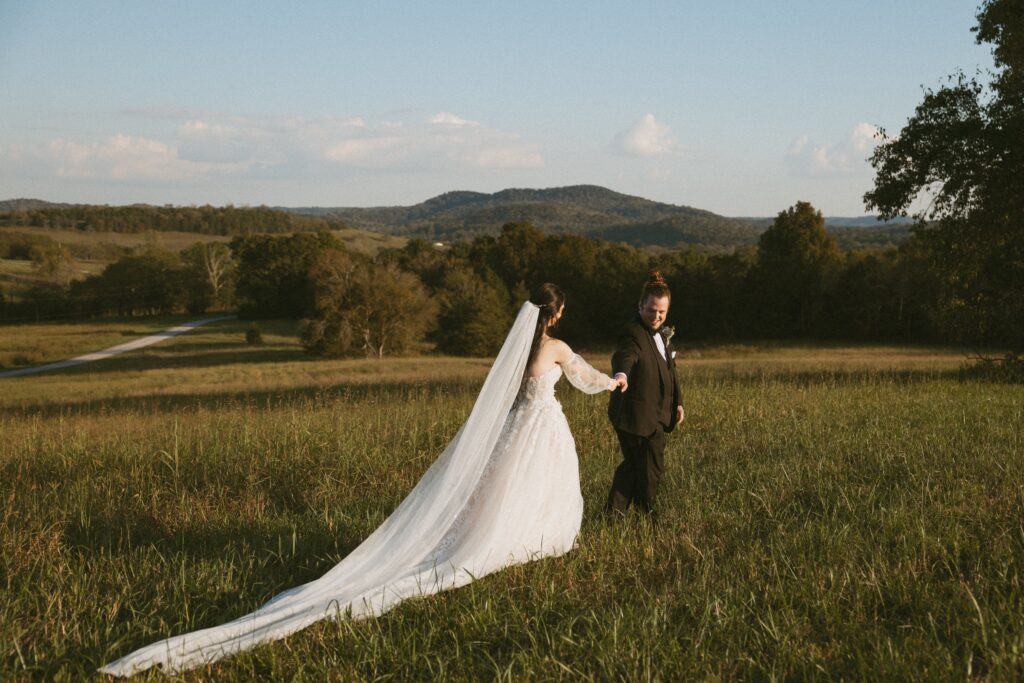 Groom leading bride through field with mountains behind them.