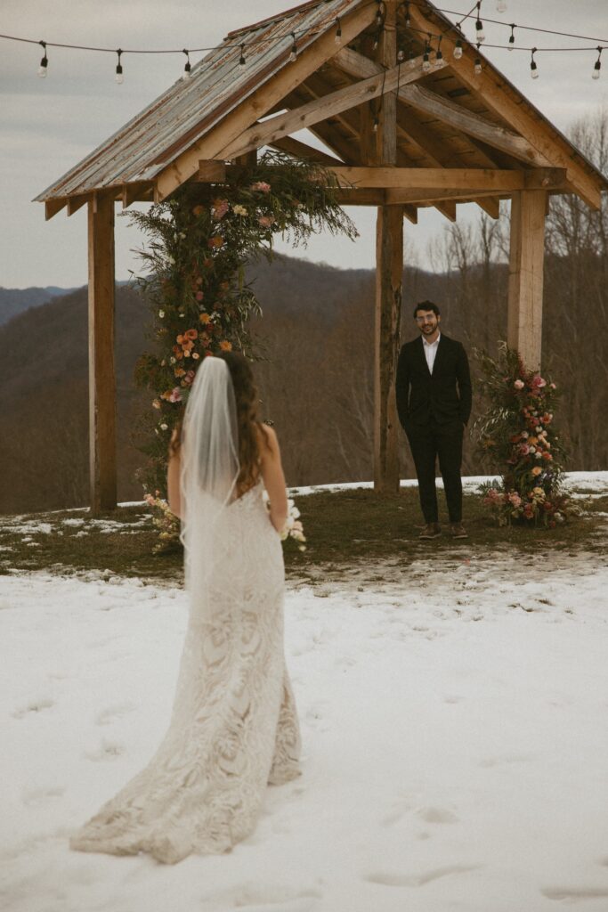 Woman in wedding dress walking towards man in suit who is standing under arch covered in flowers.