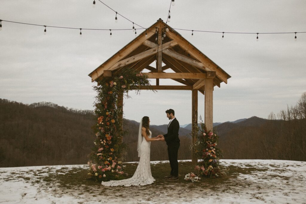 Man and woman in wedding attire standing under arch covered in flowers, holding hands and looking at each other, with mountains behind them.