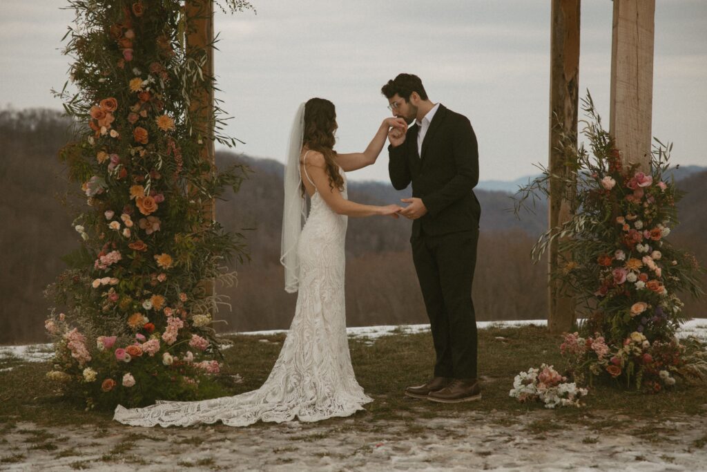 Man in suit kissing a womans hand who is in a wedding dress with flowers around them and mountains behind them.