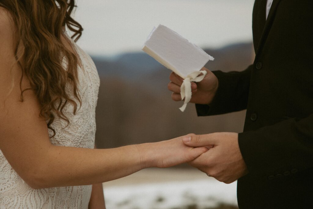 Close up of man and woman holding hands while he is reading wedding vows with a vow book. 