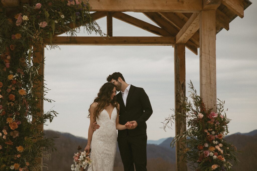 Man whispering in womans ear with flowers around them and mountains behind them.