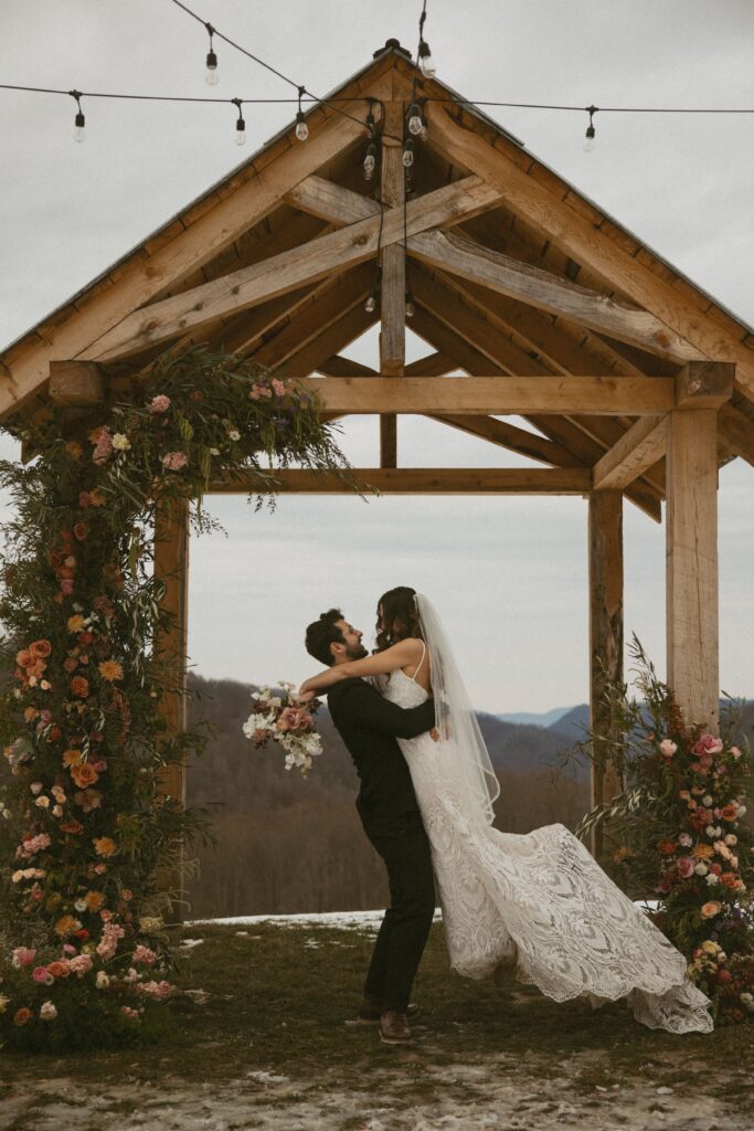 man in a suit spinning a woman in a wedding dress with flowers around them and mountains behind them.