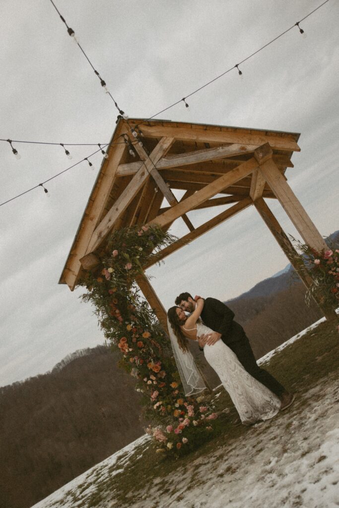 Man and woman in wedding attire hugging each other under arch covered in flowers with mountains behind them. 