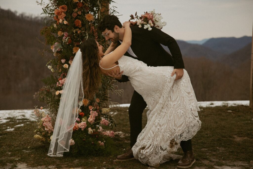 Man dipping woman in wedding dress with mountains behind them.