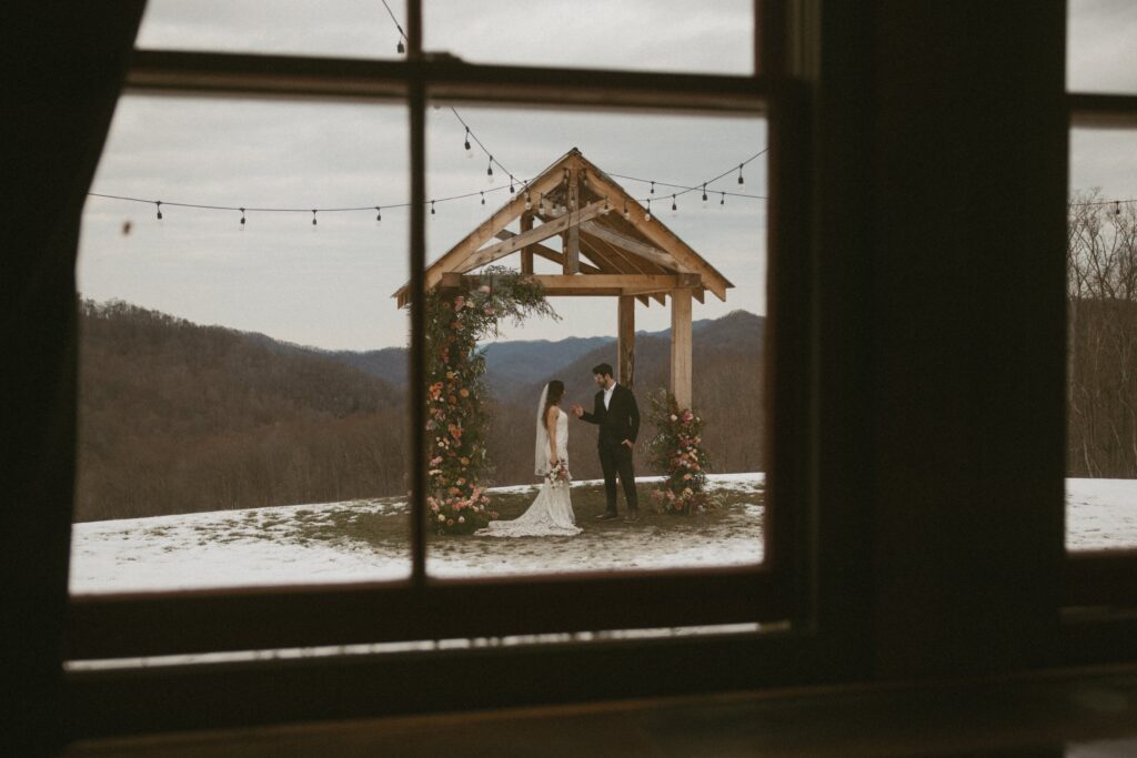 Man and woman in wedding attire through a winder standing under arch with flowers and mountains behind them.