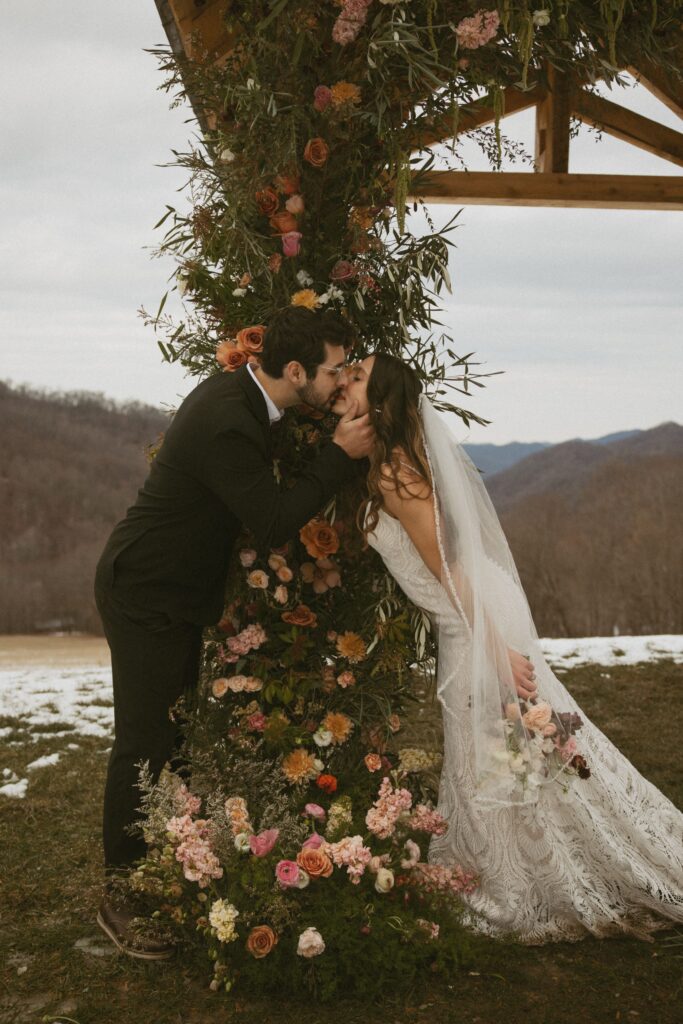 Man and woman in wedding attire kissing each other around a post covered in flowers.