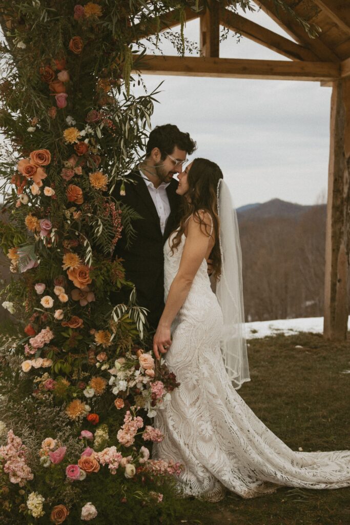 Man and woman in wedding attire leaning against a post covered in flowers.