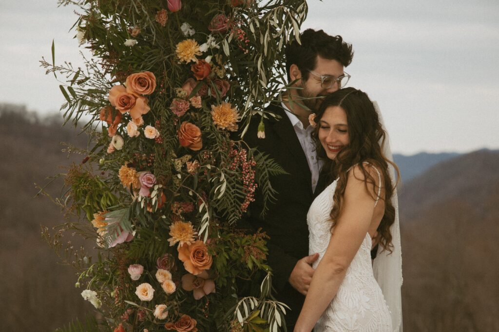 man kissing woman on the head while they are smiling and leaning on a post covered in flowers with mountains behind them.