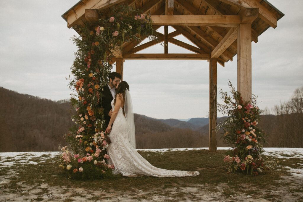 Man and woman in wedding attire leaning on arch way covered in flowers with mountains behind them. 