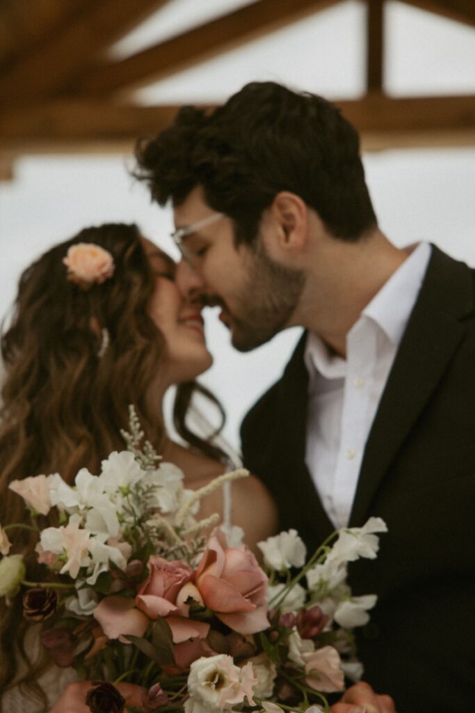 Man and woman in wedding attire going in for a kiss with bouquet in foreground. 