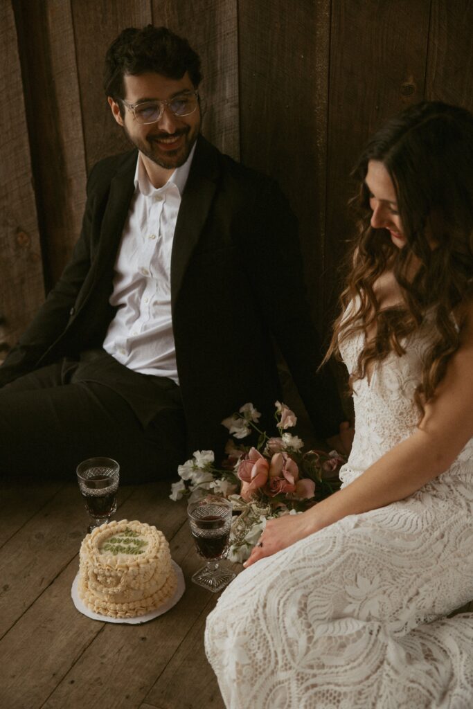 Man and woman in wedding attire sitting on porch smiling with cake between them.