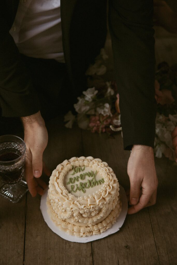 Man in suit picking up a small elopement cake that says "in every lifetime".