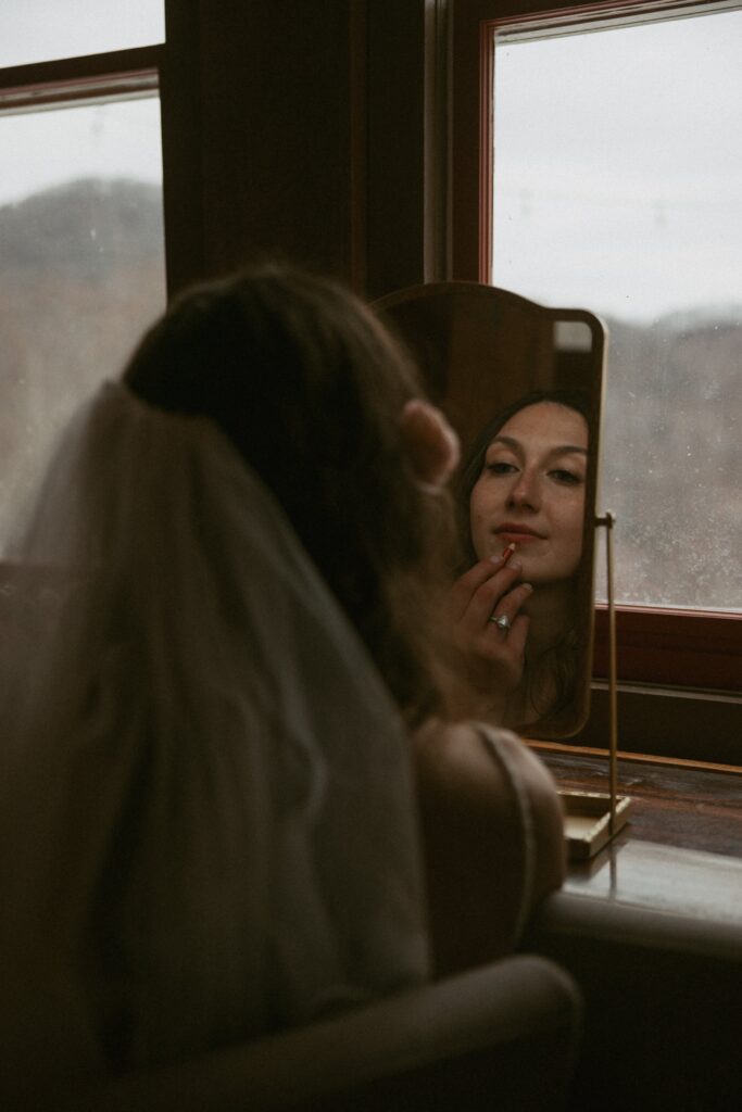 woman in wedding attire looking in a mirror touching up lipstick with mountains behind her.