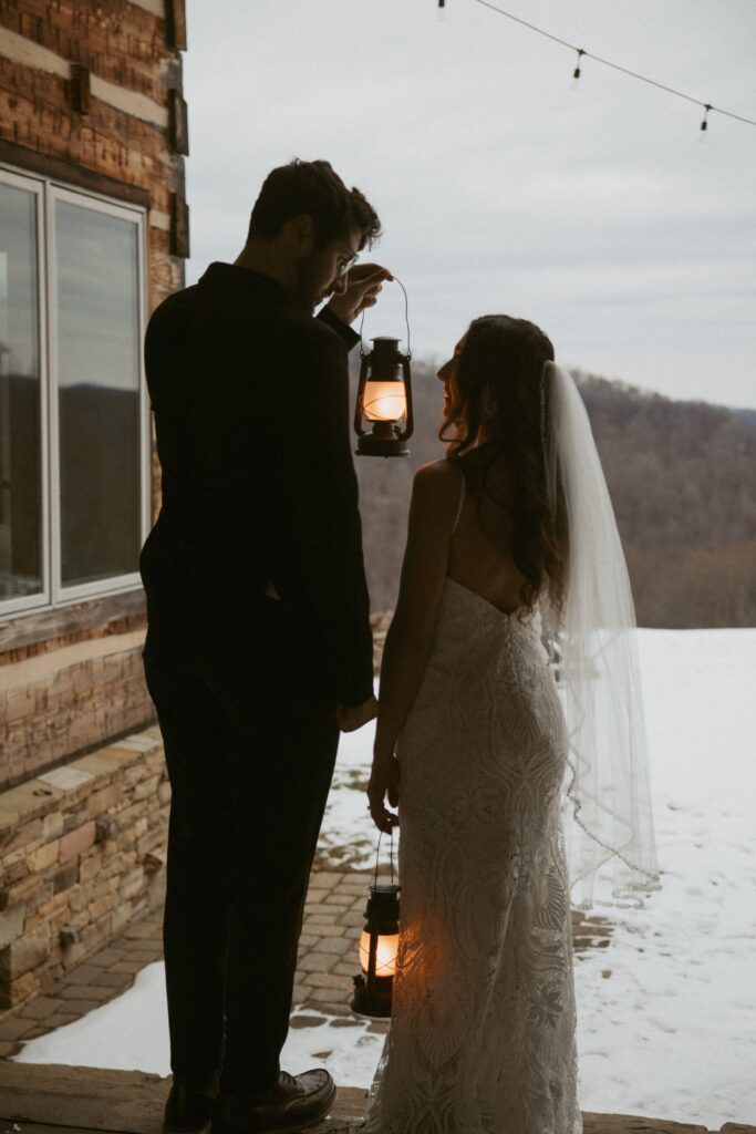 Man and woman in wedding attire holding lanterns on cabin porch. 