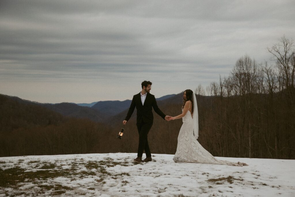 Man and woman in wedding attire walking in the snow with a lantern and mountains behind them.