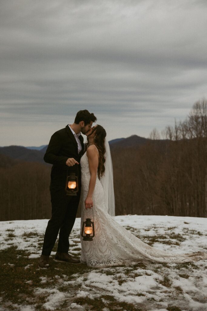 Man and woman in wedding attire holding lanterns and kissing with mountains behind them.