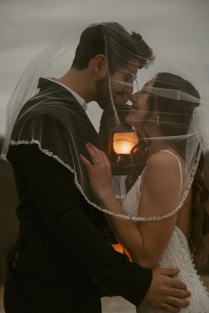 Man and woman in wedding attire nose to nose underneath veil while holding lanterns.