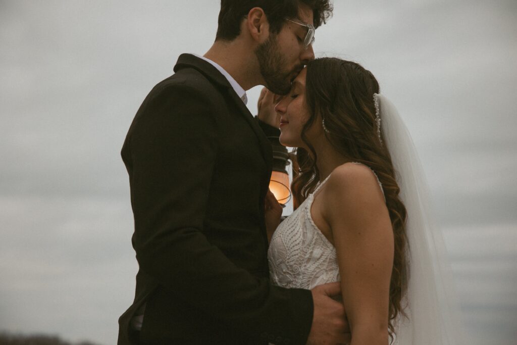 Man in suit and holding a lantern kissing a woman in wedding dress on the forehead.