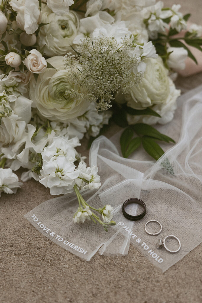 White floral bouquet laying with sheer gloves and wedding rings on the ground.